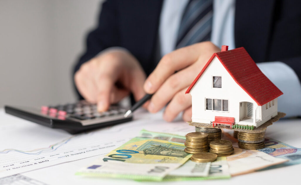 Small model house resting on stacked coins and cash beside a calculator, representing real estate financing and loan calculations.